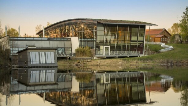 Un edificio moderno con fachada de vidrio, un reflejo en el agua y una estructura pequeña adyacente.
