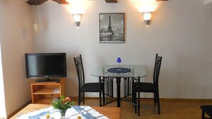 Un comedor con mesa de vidrio, sillas negras, un televisor sobre un soporte de madera y un cuadro con vista a un paisaje urbano.