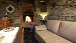 Une pièce aux murs de pierre, avec une cheminée, un canapé, une horloge et une petite table sur laquelle se trouvent une bouteille et des verres.