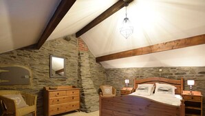 Une chambre avec un mur en pierre, un lit en bois, une commode, une chaise en osier et un luminaire suspendu.