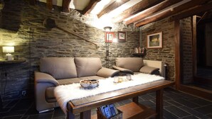 Une pièce chaleureuse avec des murs en pierre, des poutres apparentes, un canapé, une table recouverte d’un plaid en fourrure et un lampadaire.