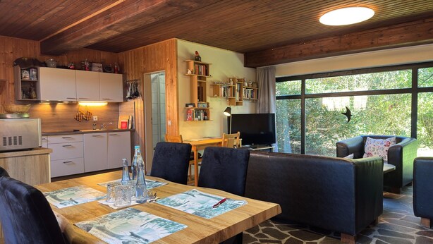 A cozy interior with a wooden ceiling, a dining area with placemats, a TV, and a view of greenery outside. - Apartment Manderscheid Near Castle Ruins (Manderscheid)
