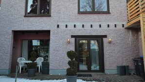 A two-story building with a pink textured exterior, a front door with glass panels, and a small patio area with chairs and potted plants. - Apartment Manderscheid Near Castle Ruins (Manderscheid)