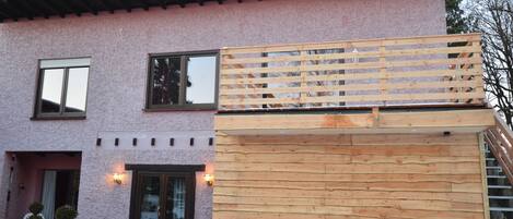 Een woonhuis met twee verdiepingen, met een roze gevel, een houten terras en een voordeur met ruitpanelen.
