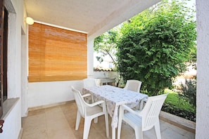 Um pátio externo com mesa e cadeiras, uma janela com persianas e um arbusto verde grande.