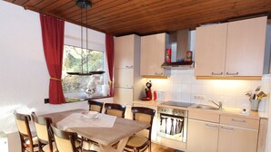 Cocina compacta con armarios de madera, una mesa de comedor con sillas y un ventanal con cortinas rojas.