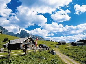 En traditionell timmerstuga med ett sluttande tak, omgiven av ett trästaket, och med berg och en klarblå himmel som bakgrund.