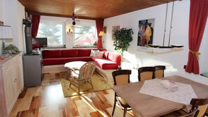 Un salón acogedor con un sofá rojo, una mesa de comedor de madera y sillas. Hay una ventana con cortinas, una planta y un reloj de pared.