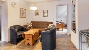 A living room with a brown sofa, two armchairs, a wooden coffee table, and a hanging light fixture. There are framed pictures on the wall and a clock.