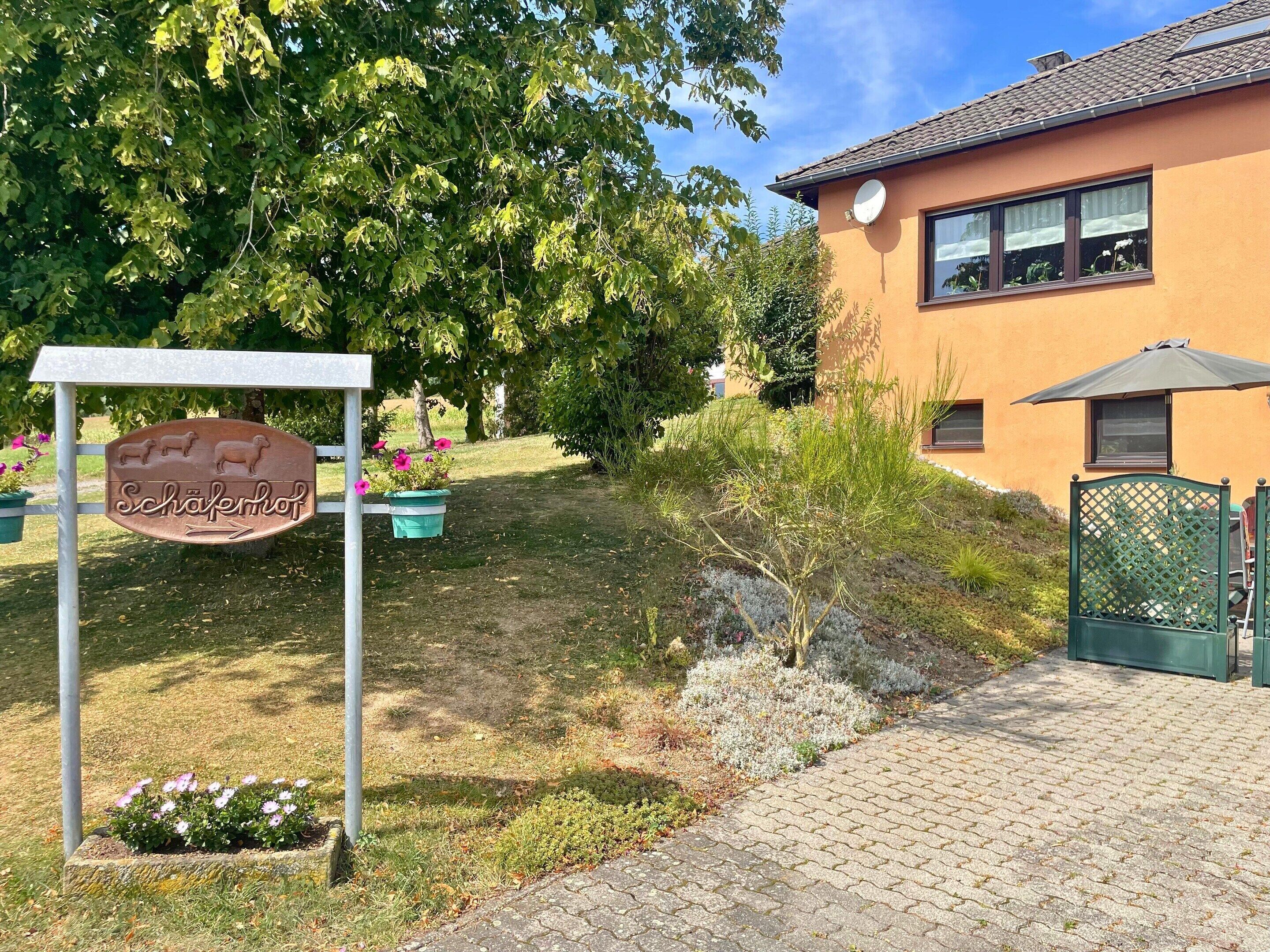 A sign for 'Schäferhof' with a garden area, a building with a tiled roof, and a paved walkway.