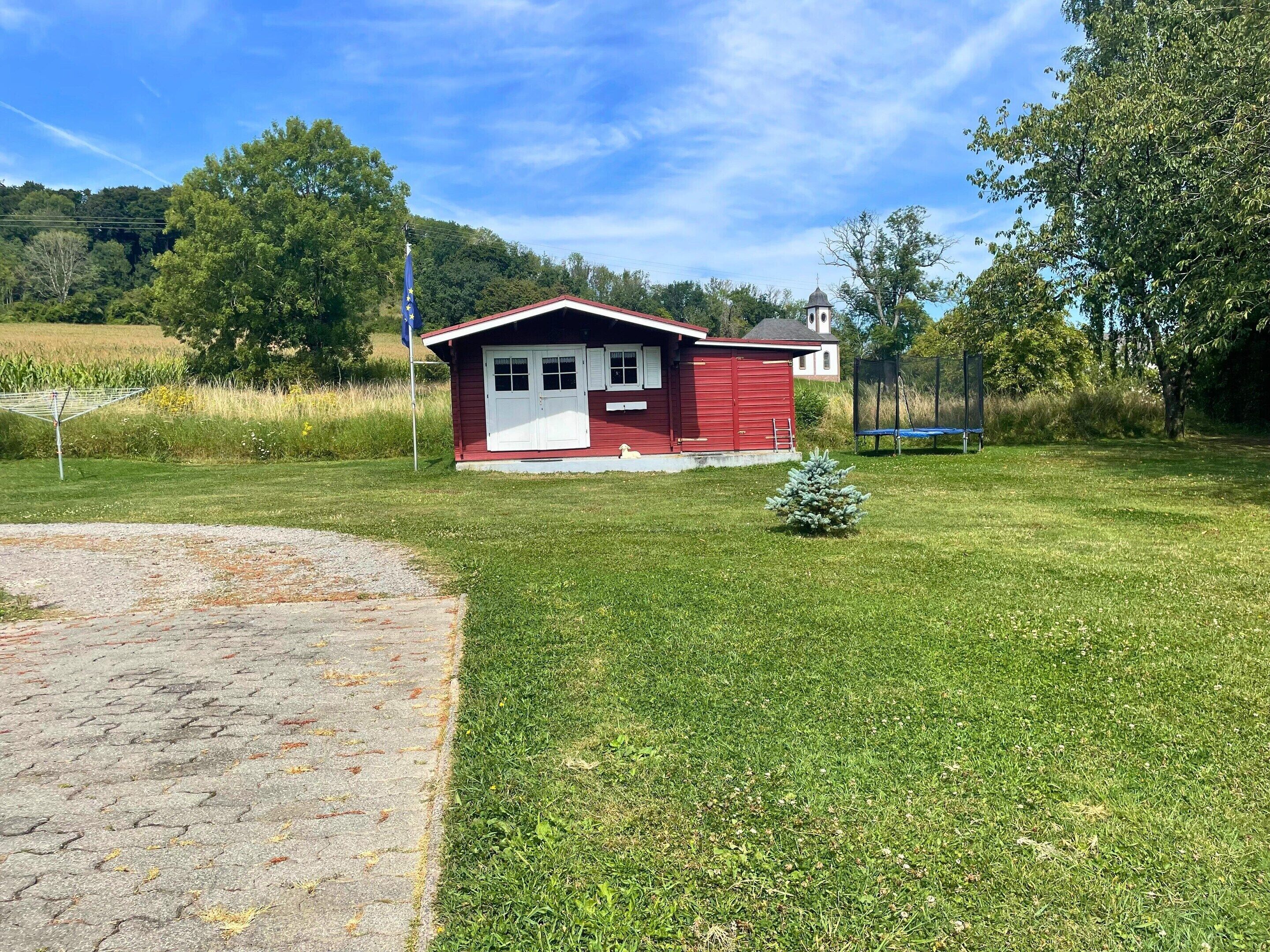 A small red building with a white door and windows, surrounded by a grassy area with a small tree and a trampoline.