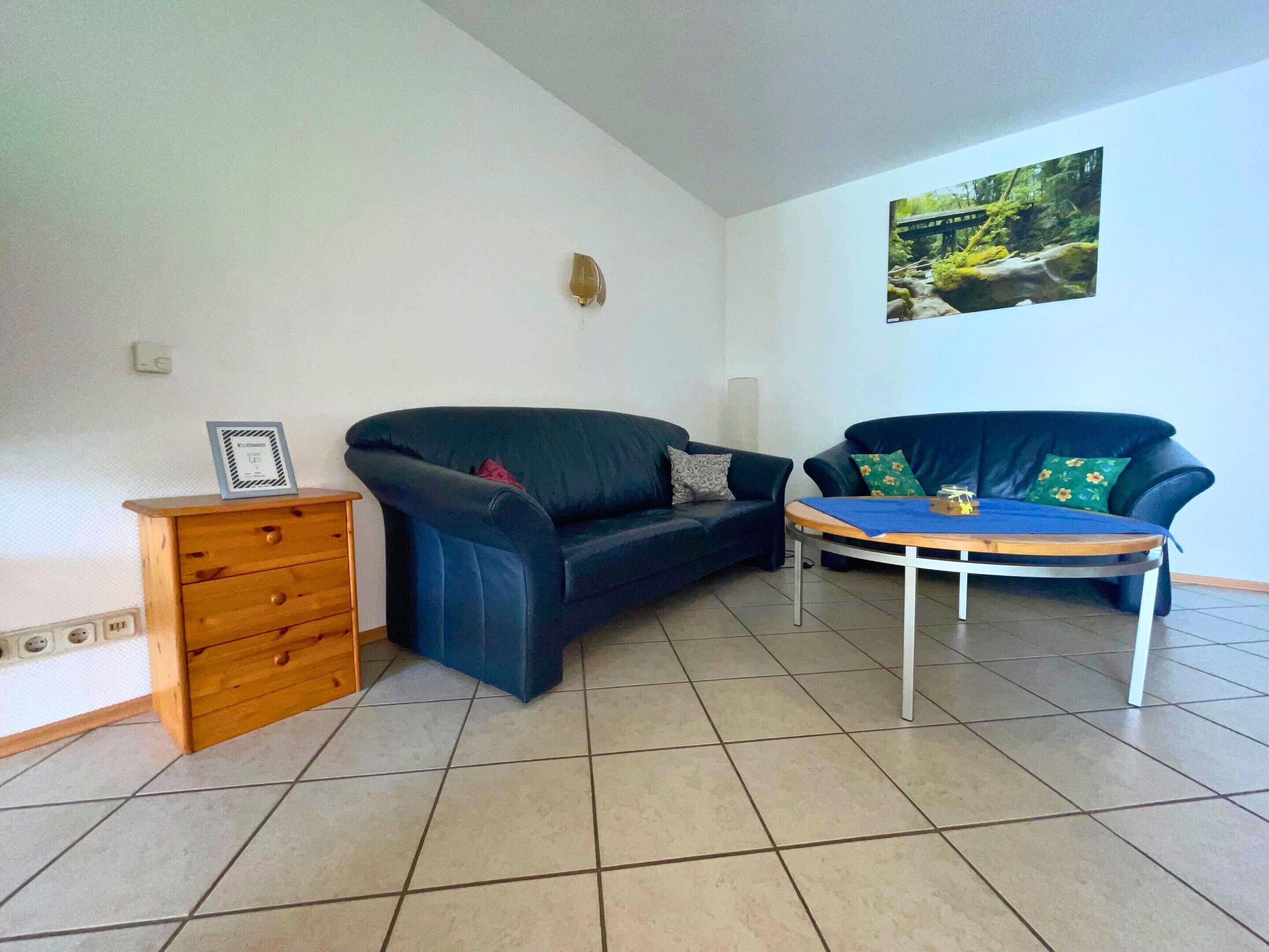 A living room with two dark blue sofas, a wooden side table, a tiled floor, and a framed picture on the wall.