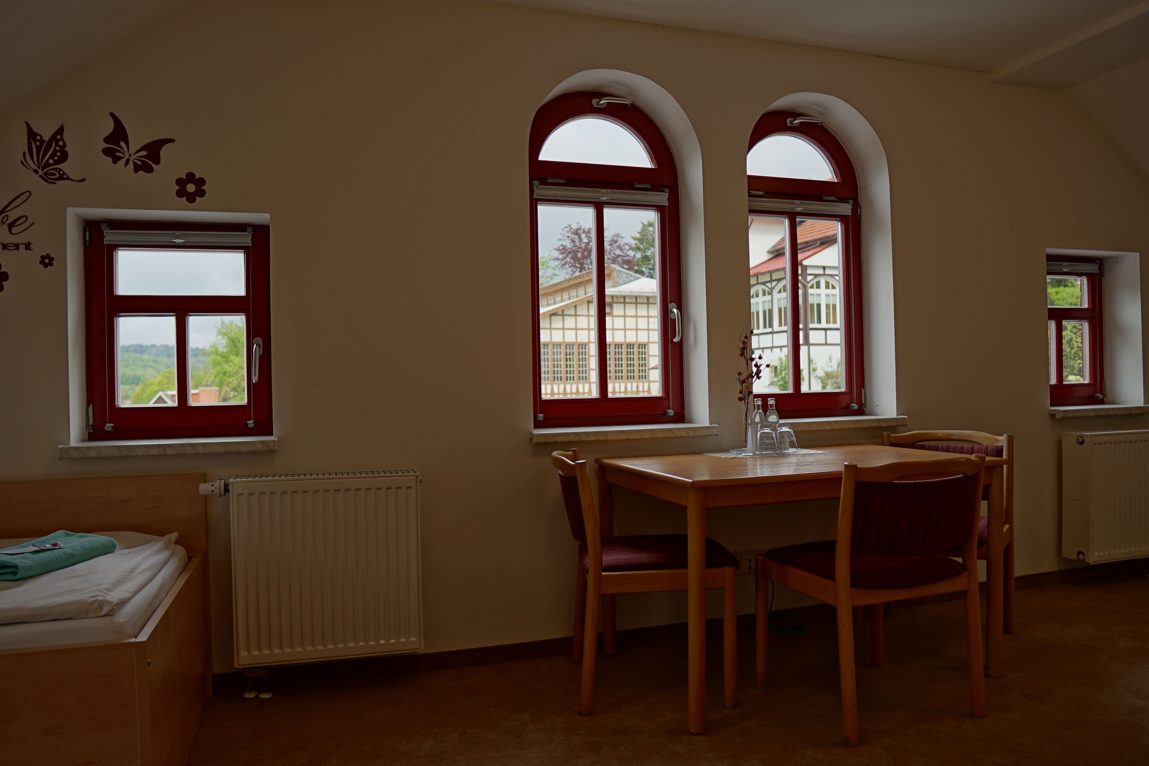 A room with a wooden bed, a dining table with chairs, and two arched windows with red frames.