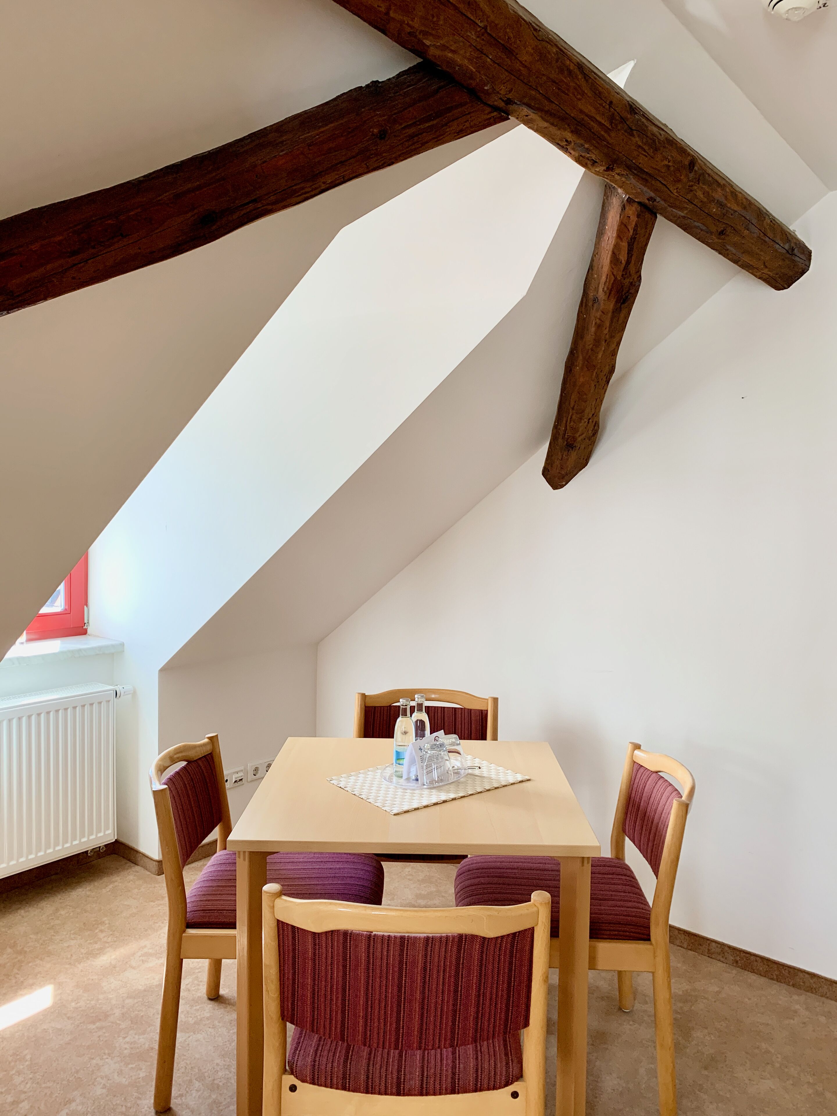 A dining area with a wooden table and four chairs, a radiator, and a sloped ceiling with exposed beams.