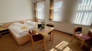 Een hotelkamer met een bed, twee nachtkastjes, een houten stoel, een kleine tafel met een stoel, en een raam met gordijnen.