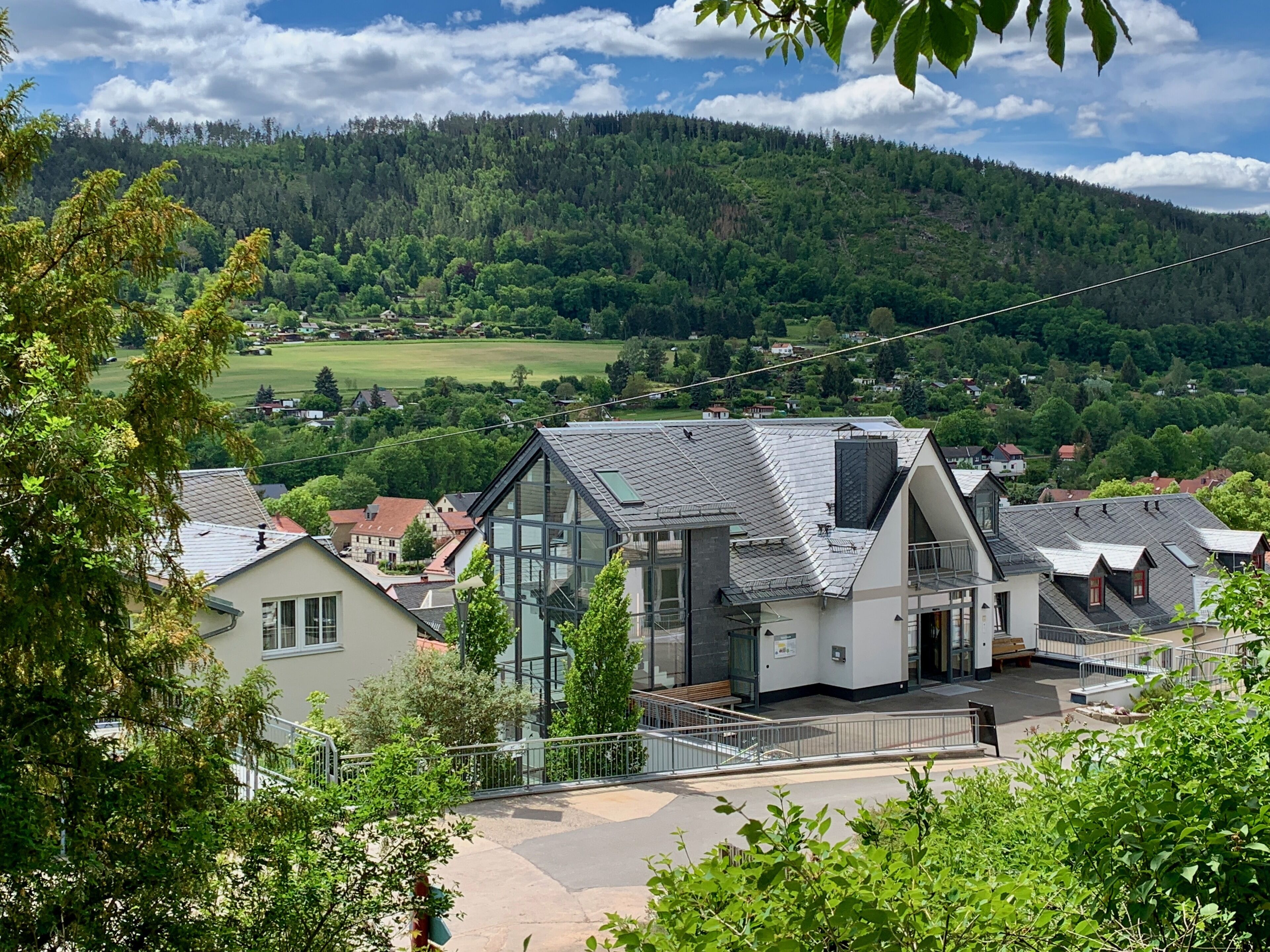 Ein modernes Gebäude mit großen Fenstern und einem geneigten Dach, umgeben von üppiger Vegetation und einer Bergkulisse.