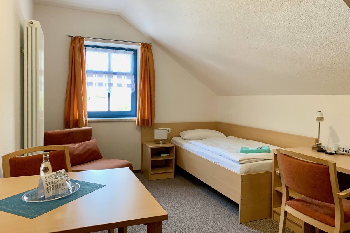 A small, neatly arranged room with a bed, a desk, a chair, a table, and a window with curtains.