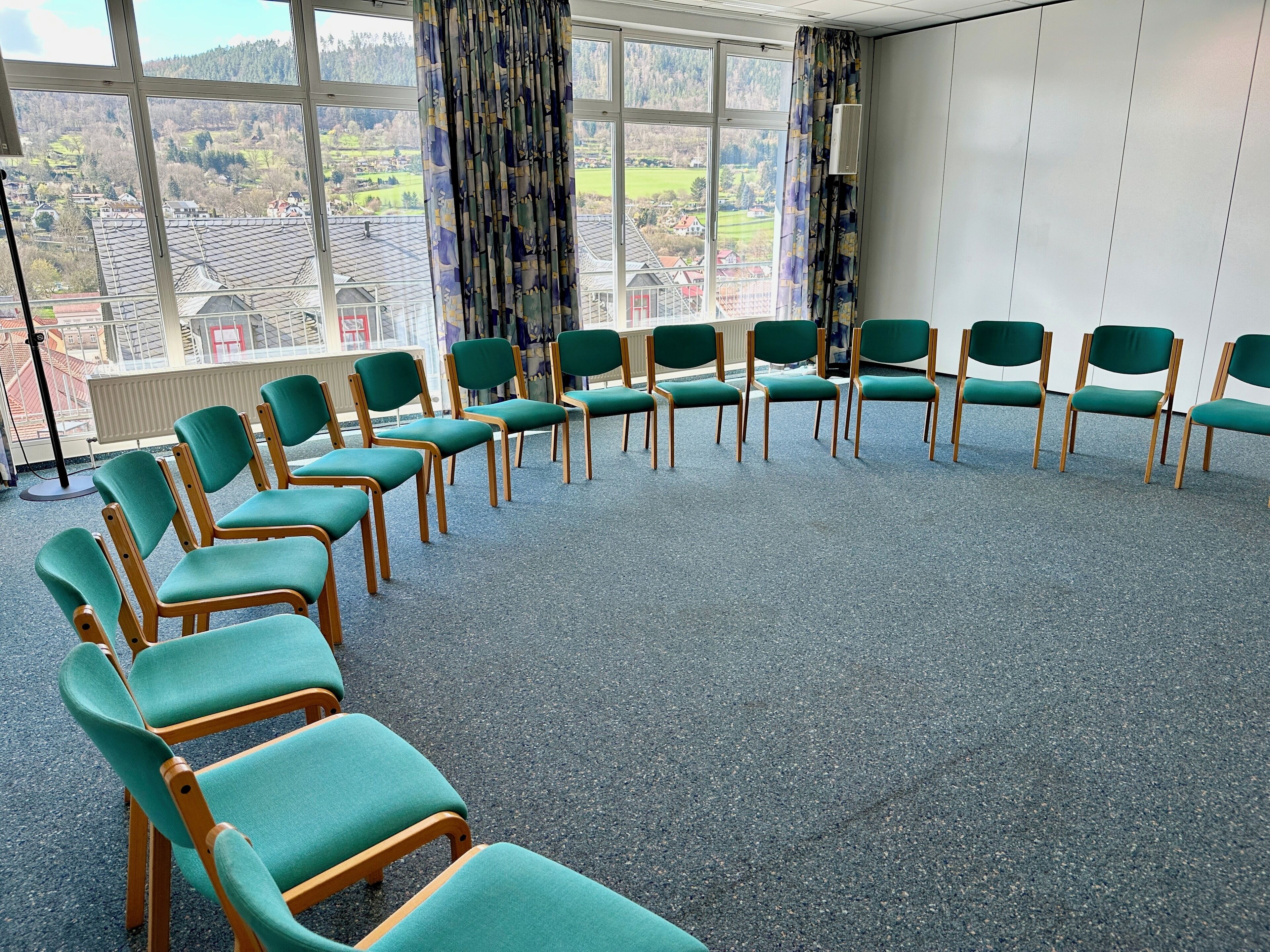 Un salón de conferencias con sillas verdes dispuestas en semicírculo, amplias ventanas con vistas a un paisaje montañoso y un radiador en la pared.