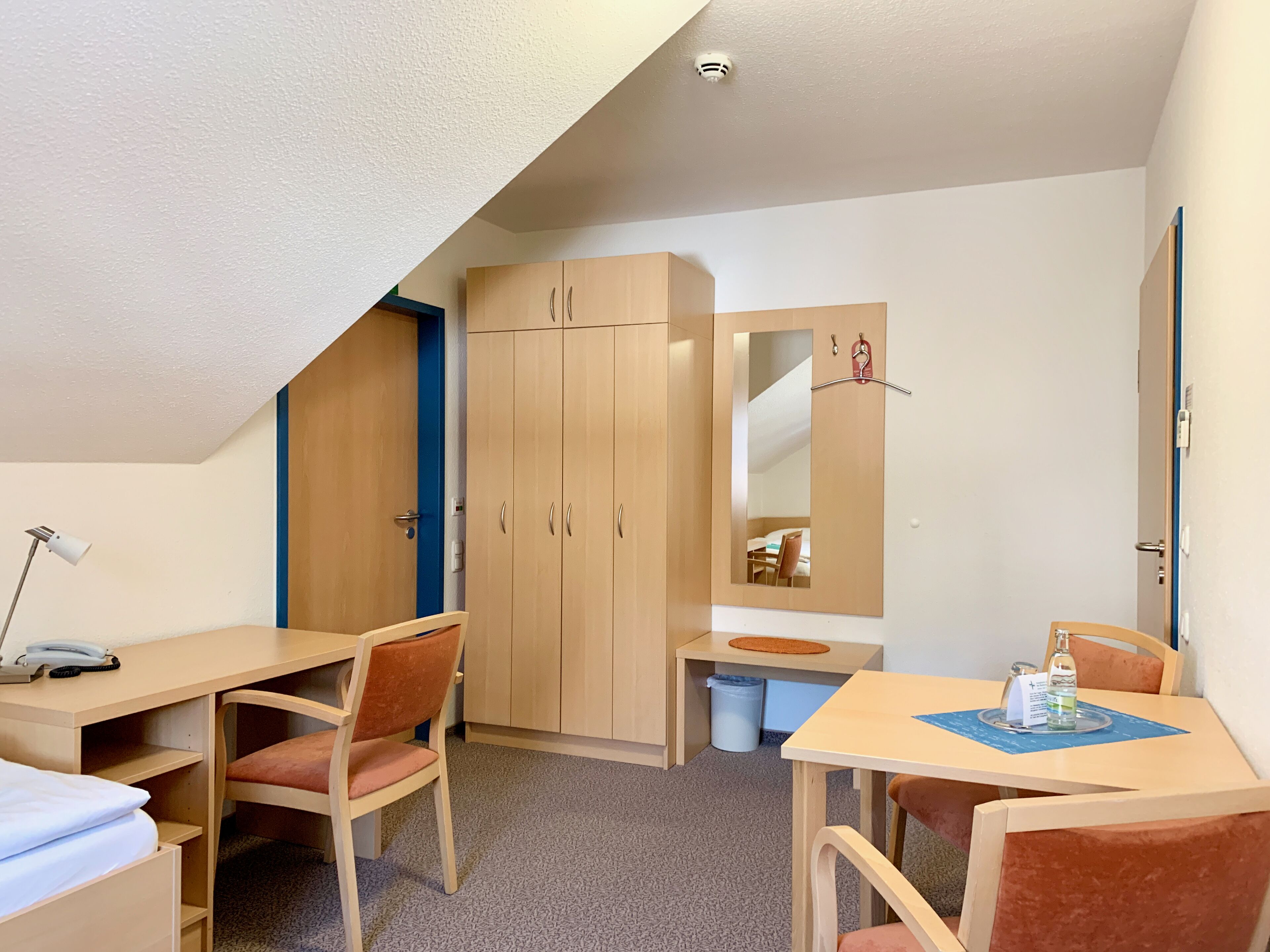 A compact hotel room with a single bed, a desk, a chair, a small table with a bottle, and a mirror.