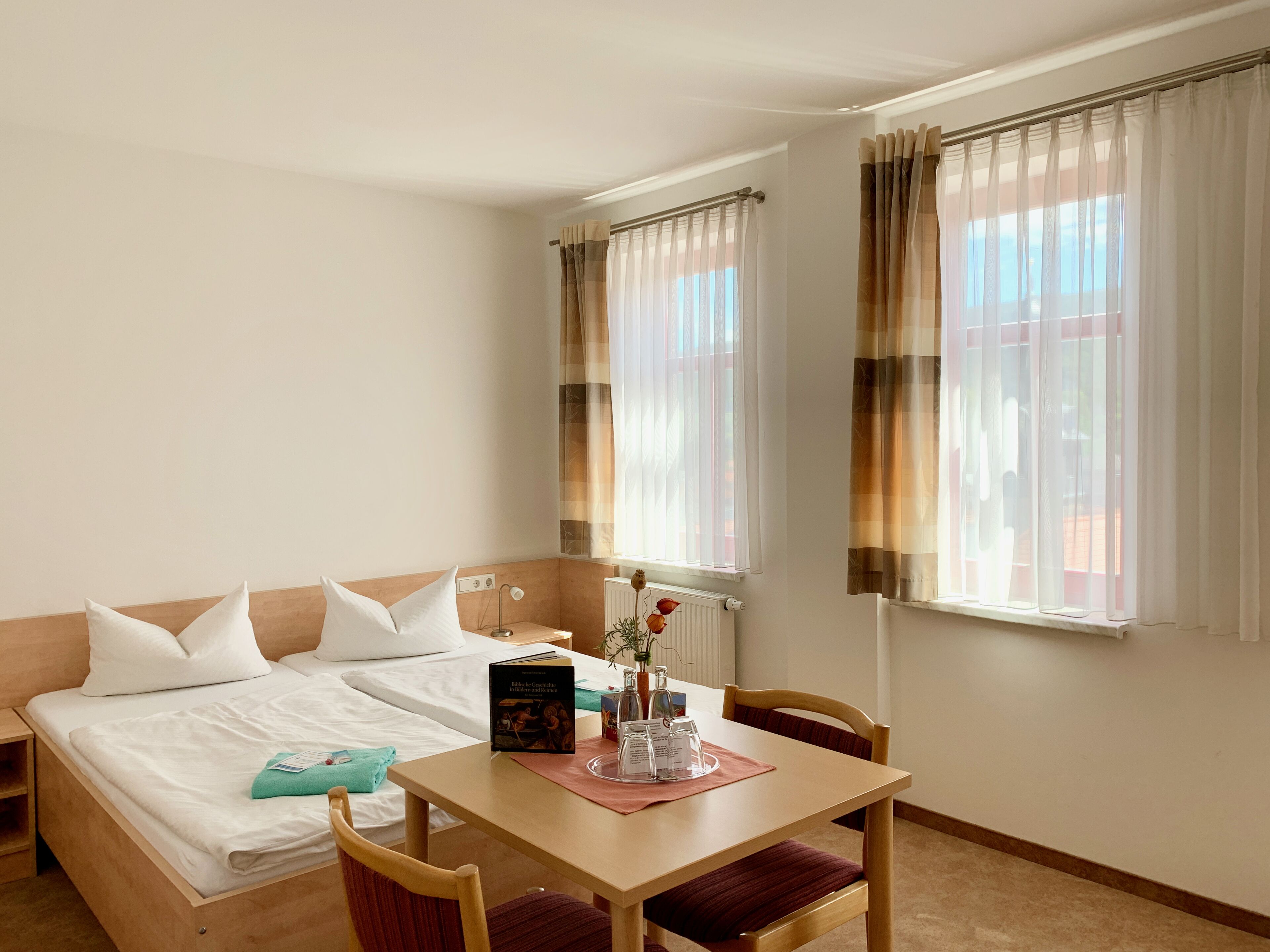 Una habitación de hotel con una cama, una mesa con un libro y un vaso, y dos ventanas con cortinas.