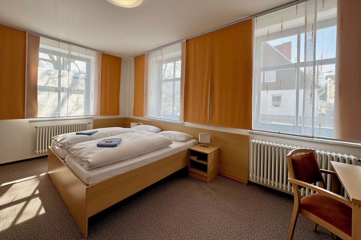 A hotel room with a bed, a small bedside table, a chair, and large windows with curtains.