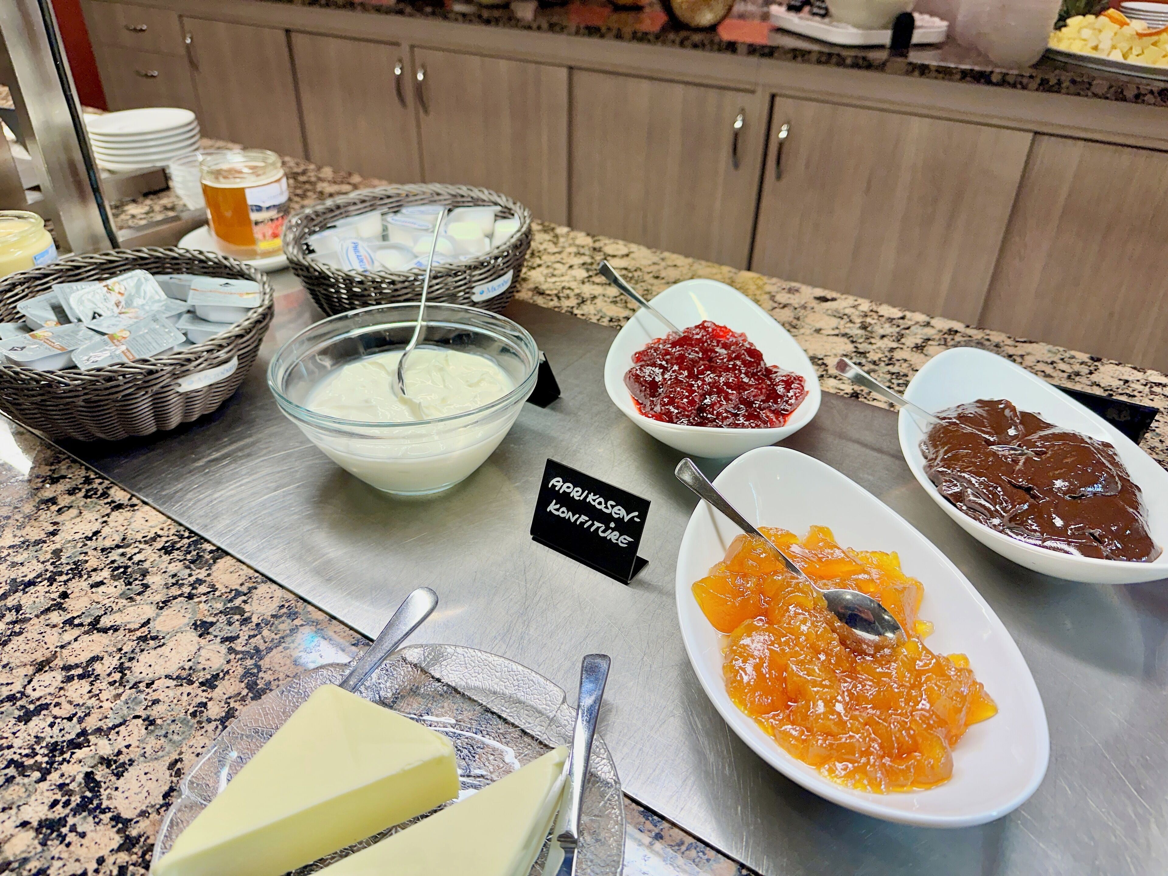Une table de buffet proposant divers aliments, notamment du yaourt, de la compote de fruits et une pâte à tartiner au chocolat.