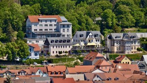 Eine Wohngegend an einem Hang mit verschiedenen Häusern, darunter ein großes, zweistöckiges Gebäude mit rotem Dach und ein kleineres, angrenzendes Gebäude mit grauem Dach.