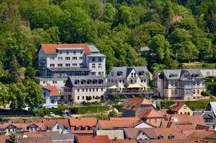 Uma área residencial em encosta com diversas casas, incluindo um edifício grande de dois andares com telhado vermelho e um prédio menor adjacente com telhado cinza.