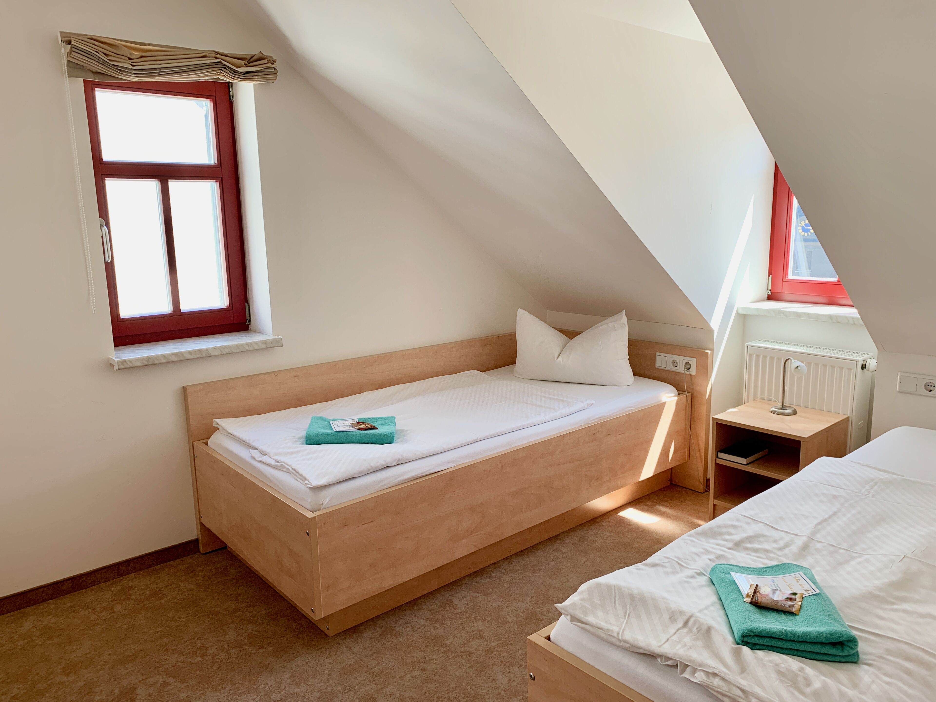 Une petite chambre avec un lit simple, un plafond en pente, une fenêtre à cadre rouge, un radiateur et une table de chevet avec une lampe.