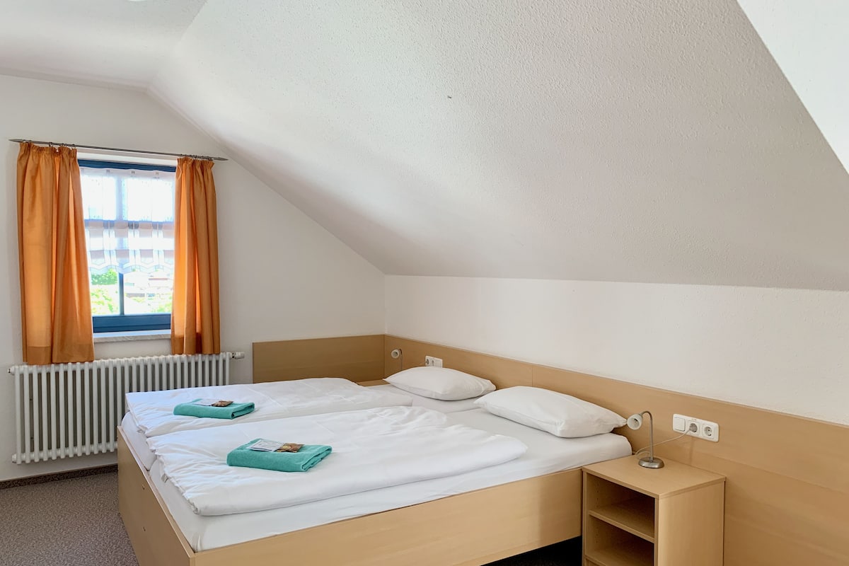 A single bed with white linens and a wooden headboard, a bedside table with a lamp, and a window with orange curtains.