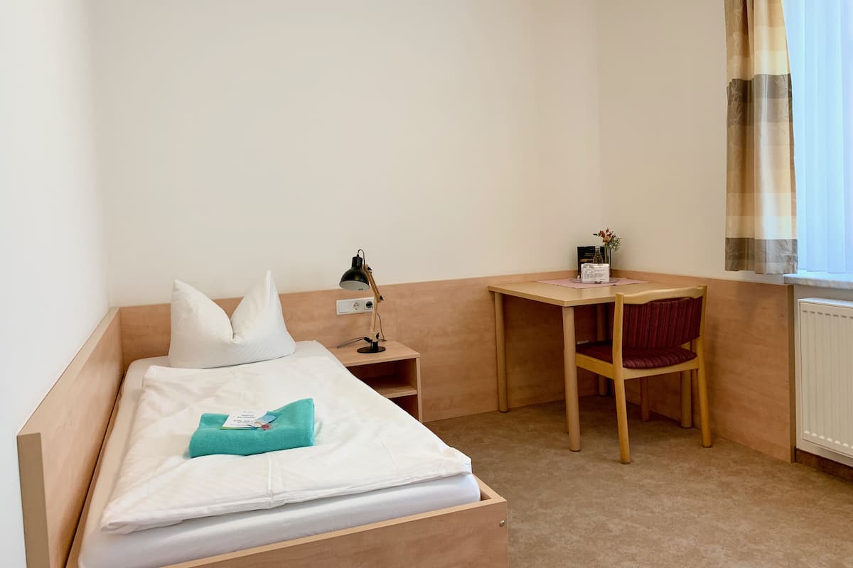 A hotel room with a bed, a desk, a chair, a lamp, a window with curtains, and a radiator.