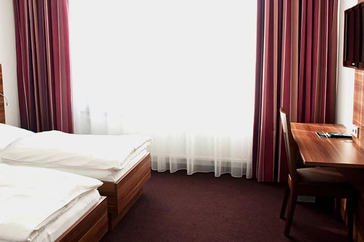 A hotel room with two wooden beds, a desk with a chair, a flat-screen TV, and a large window with sheer curtains.