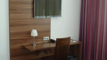 Ein Hotelzimmer mit Holzschreibtisch, einem an der Wand befestigten Fernseher, einem Sessel, einer Lampe, einem Papierkorb und einem Bett mit weißer Bettwäsche.