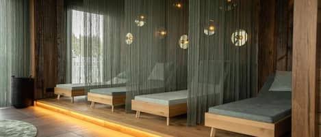 Una habitación con varias camas dispuestas junto a la pared, cortinas traslúcidas, una mesita redonda y un cubo de basura.