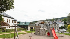 A playground with a slide and swings, surrounded by a sandy area and a wooden play structure.