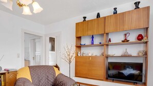 A living room with a sofa, a TV on a stand, a wooden cabinet with shelves, and a vase with branches.