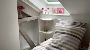 A bedroom with a sloped ceiling, a skylight, a bed with striped bedding, a bedside table with a lamp, and a shelf with decorative items. - Cottage in Central Bayeux (Bayeux)