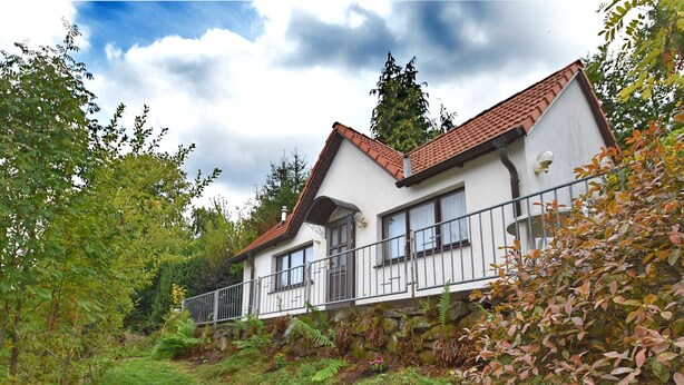 A two-story house with a white exterior and a red-tiled roof, surrounded by lush greenery and trees.