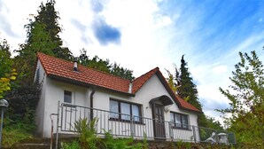 A two-story house with a red-tiled roof, white walls, and a stone foundation. There is a small porch with a metal railing and a staircase leading to the entrance.