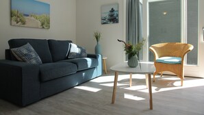 A modern living room with a dark blue sofa, a wicker chair, a small round table, and two paintings on the wall. - Stunning Apartment in Schoorl, North Holland, you can Bike to the Beach (Schoorl)