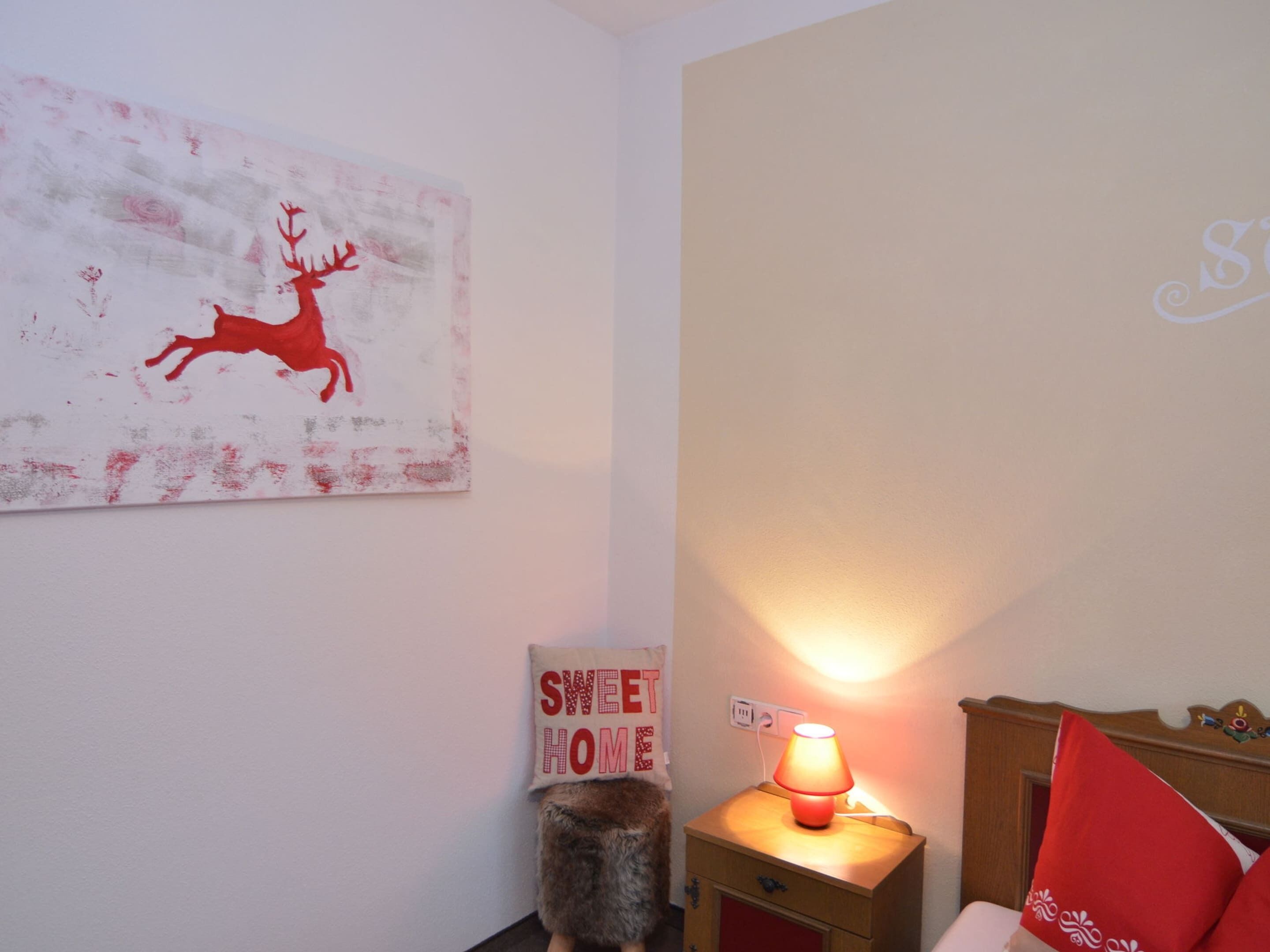 Een slaapkamer met een bed, een nachtkastje met een lamp, een zachte vloerkleden, een muur met een schilderij van een hert, en een bordje met de tekst 'sweet home'.