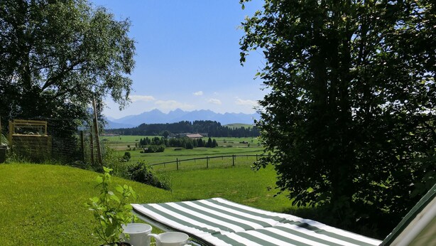 Ein grünes Areal im Freien mit einem Holztisch, der einen Blumentopf und weiße Tassen trägt, einem gestreiften Liegestuhl und Blick auf ferne Berge.