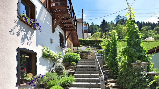 A white building with a wooden staircase leading to a balcony, surrounded by lush greenery and stone steps. - Quaint Farmhouse in Langenbach near Lake (Schleusegrund)