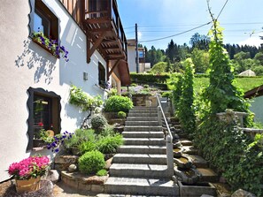 A white building with a wooden staircase leading to a balcony, surrounded by lush greenery and stone steps.