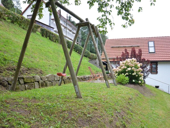 A wooden swing set in a grassy area with a stone wall and a house with a red roof in the background. - Quaint Farmhouse in Langenbach near Lake (Schleusegrund)