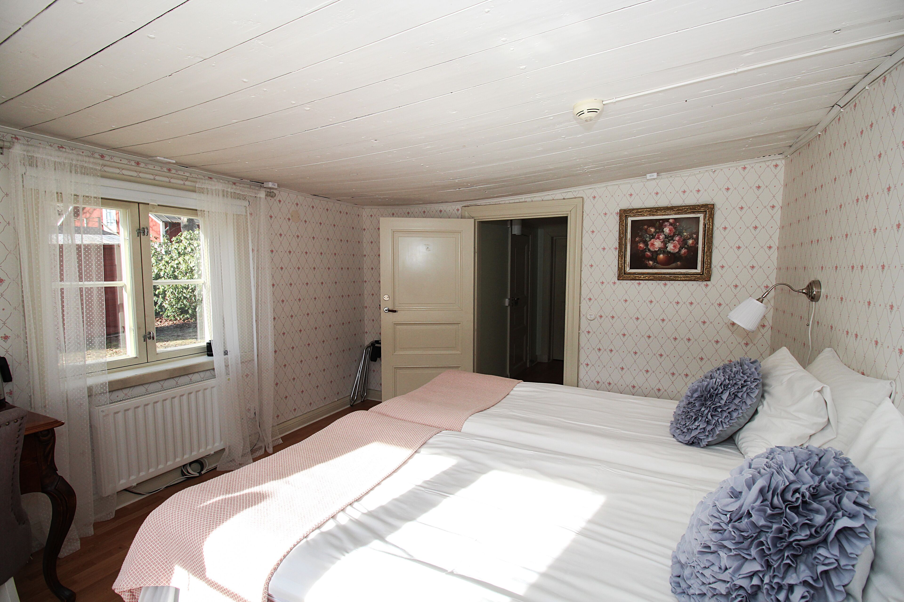 Un dormitorio con cama, un radiador, una ventana con cortinas, una puerta y un cuadro colgado en la pared.