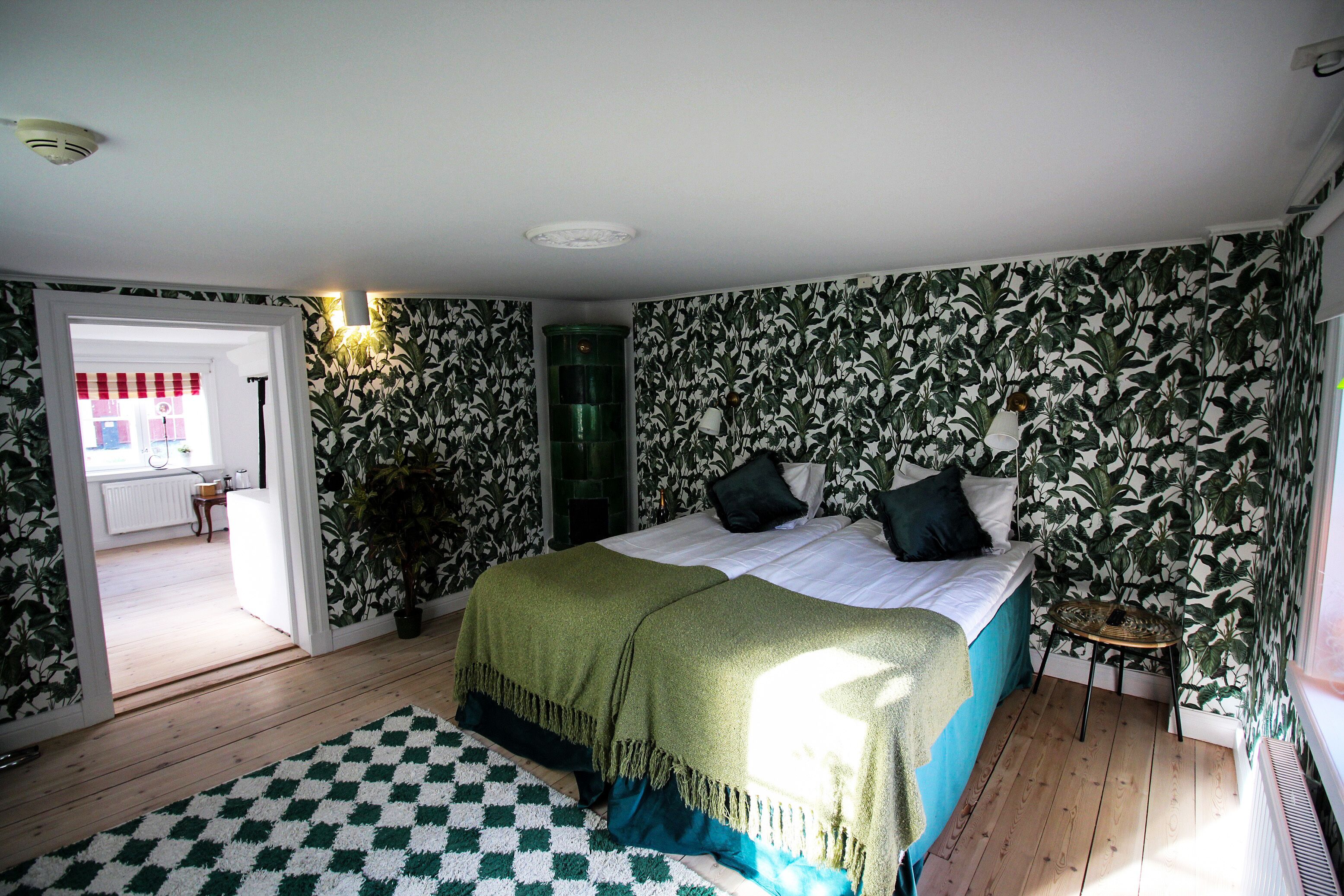 A bedroom with a bed, a green blanket, a checkered rug, and patterned wallpaper.
