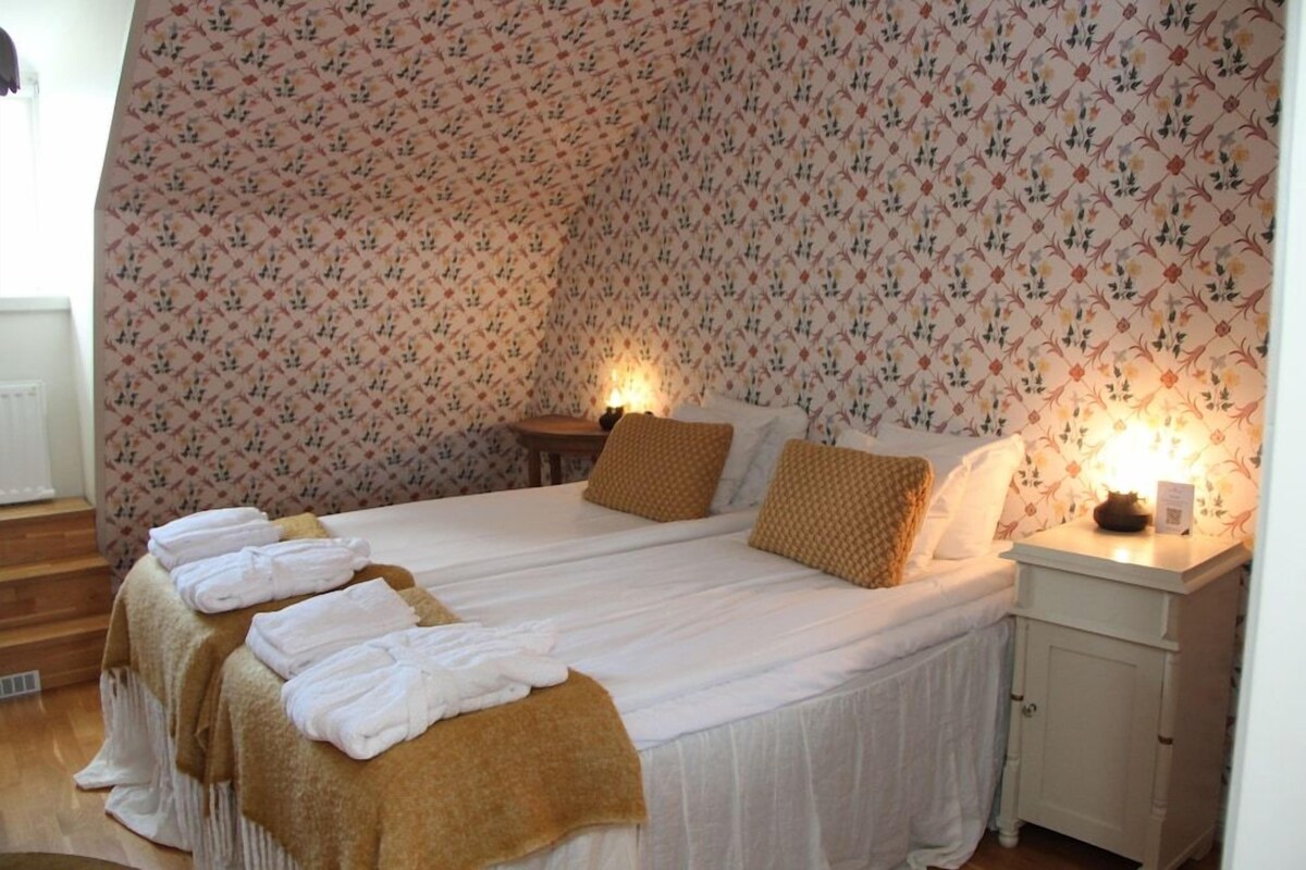 Un dormitorio con papel tapiz estampado, una cama con ropa blanca, dos mesitas de noche con lámparas y toallas sobre la cama.