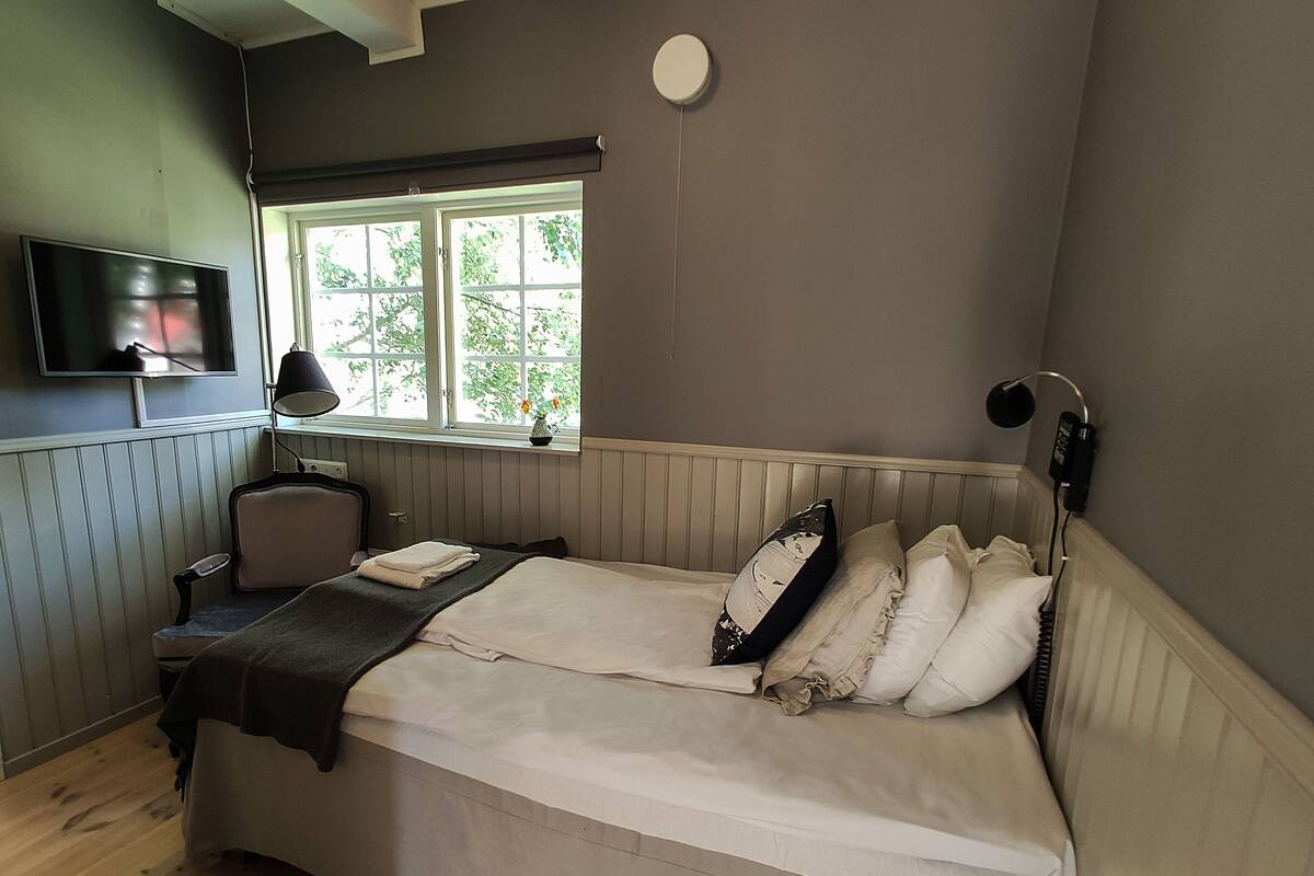 A bedroom with a bed, a desk, a chair, a TV, a window, and a lamp.