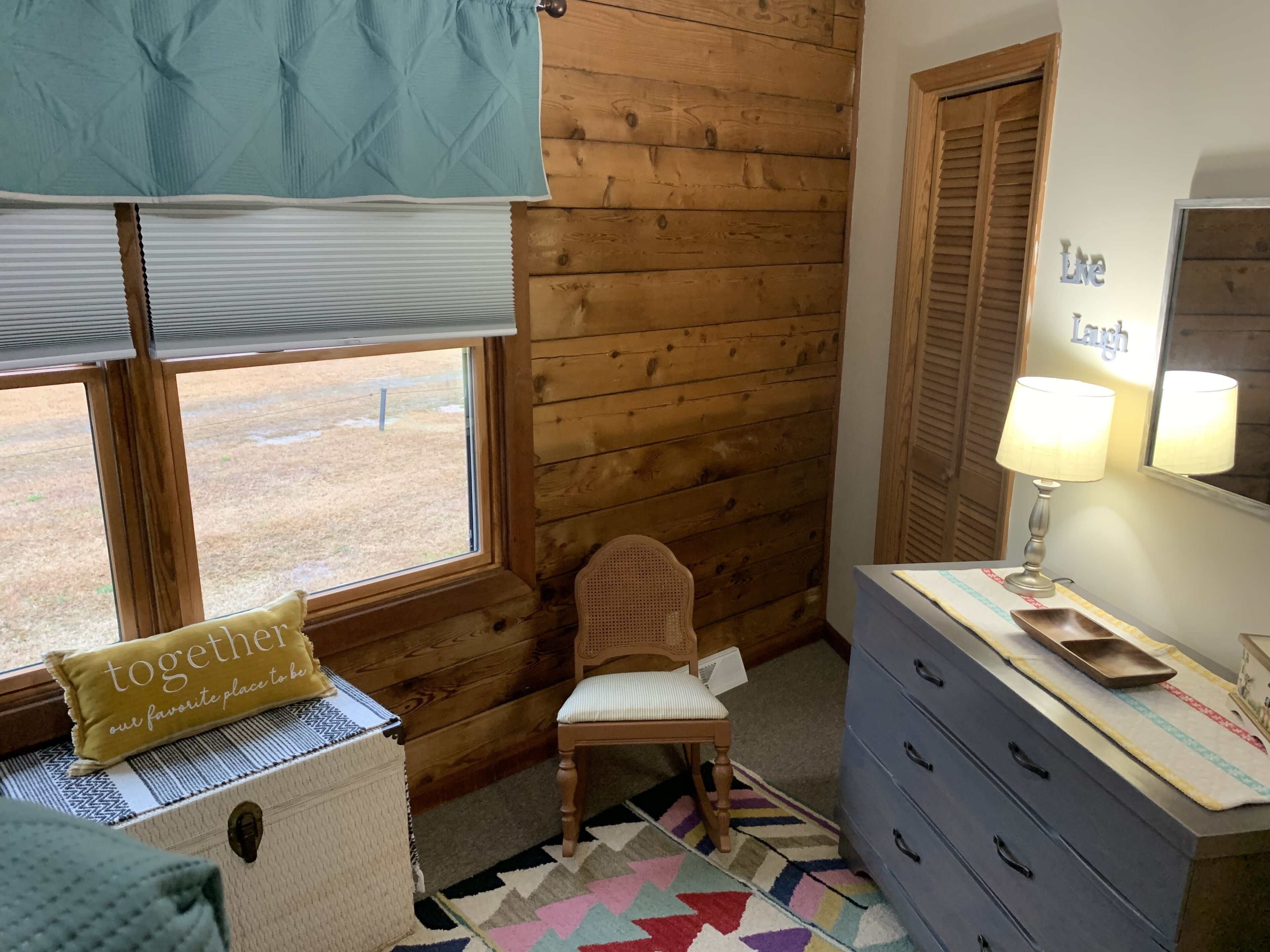A cozy room with a wooden wall, a window with blinds, a chair, a dresser with drawers, and a lamp.
